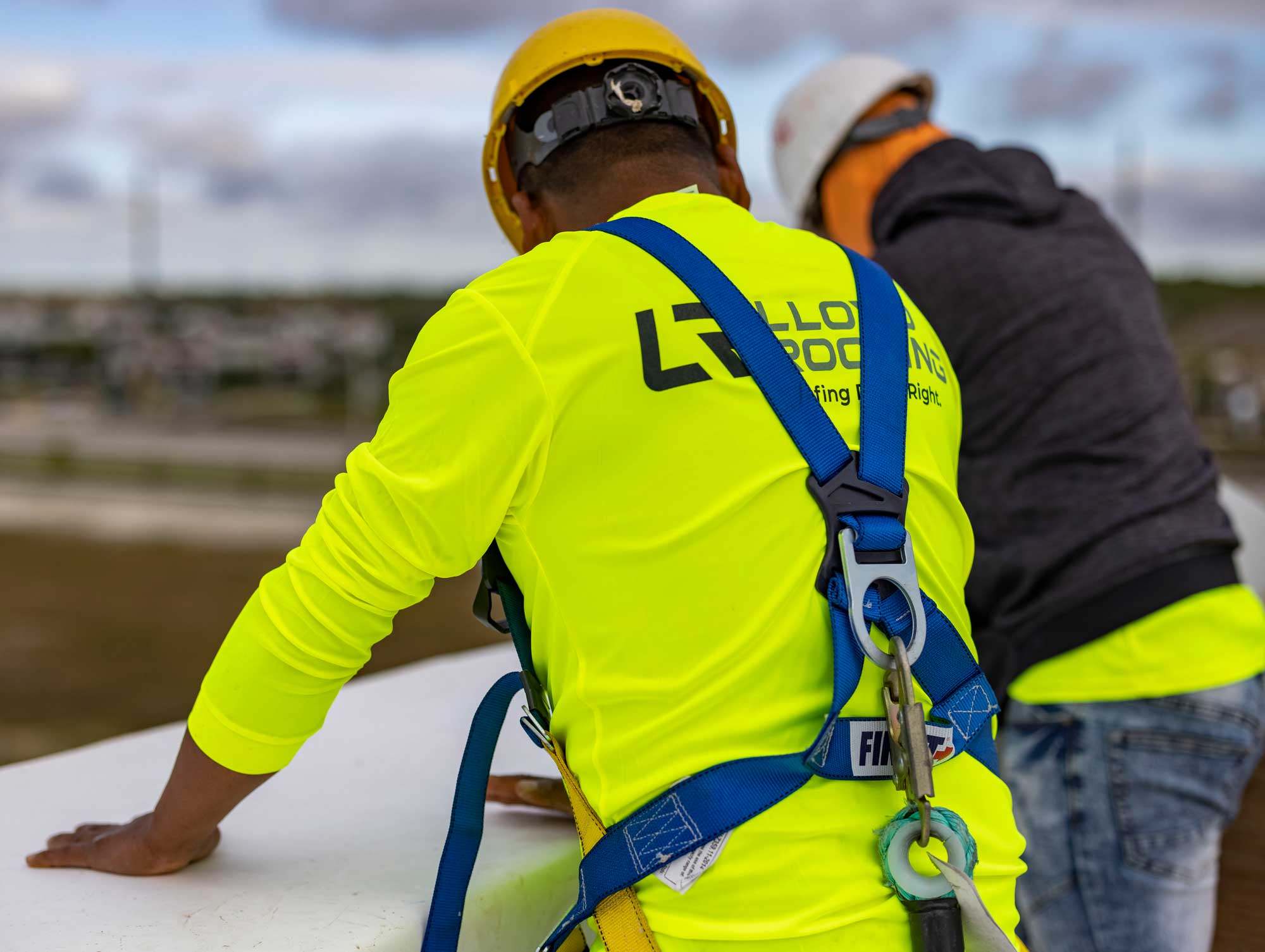 Two construction workers in safety gear work on a rooftop; one wears a yellow shirt with a company logo, safety harness, and helmet, while the other wears a white helmet and a gray jacket.